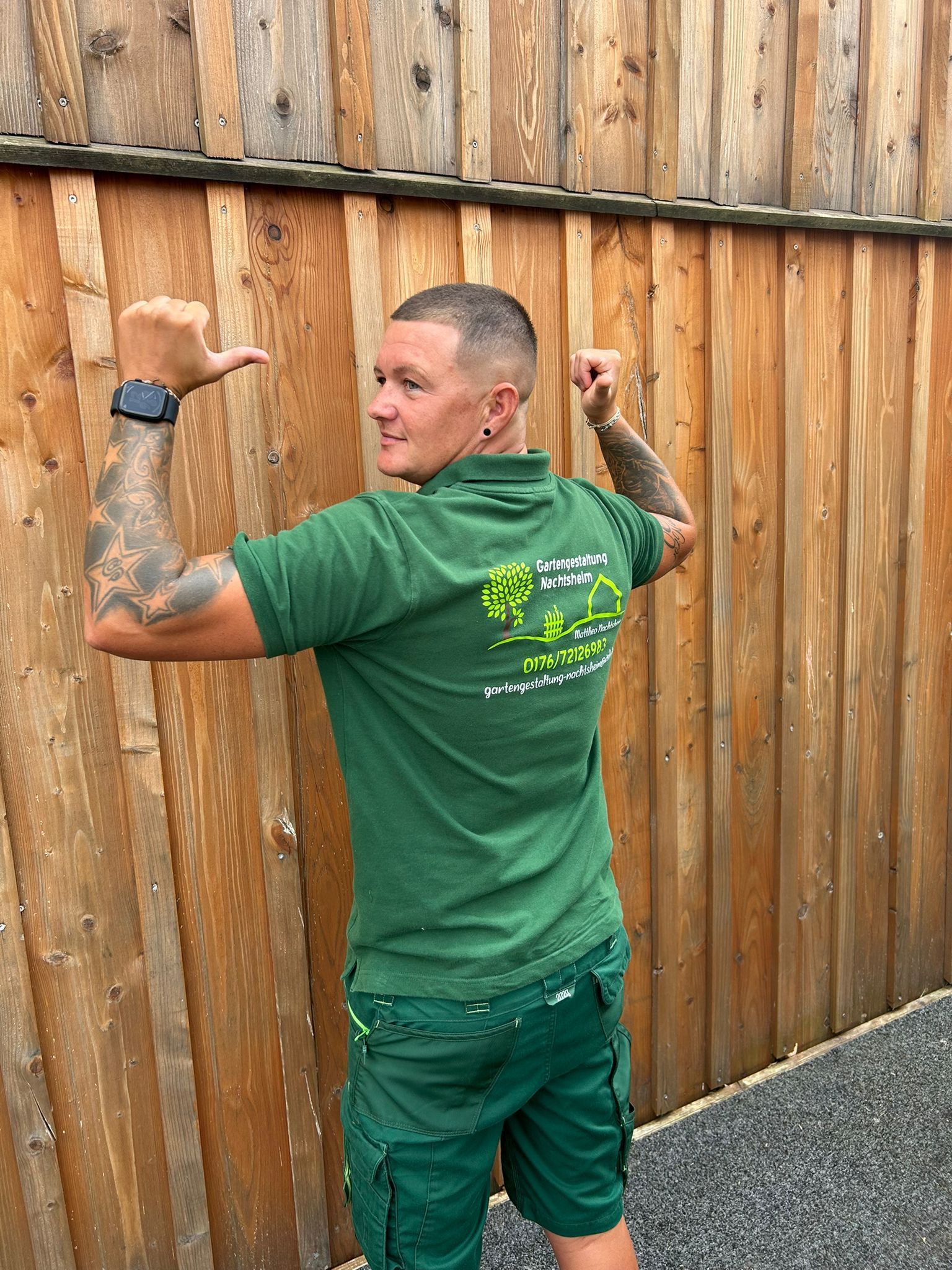 Ein Mann in einem grünen Hemd und grünen Shorts steht vor einem Holzzaun.