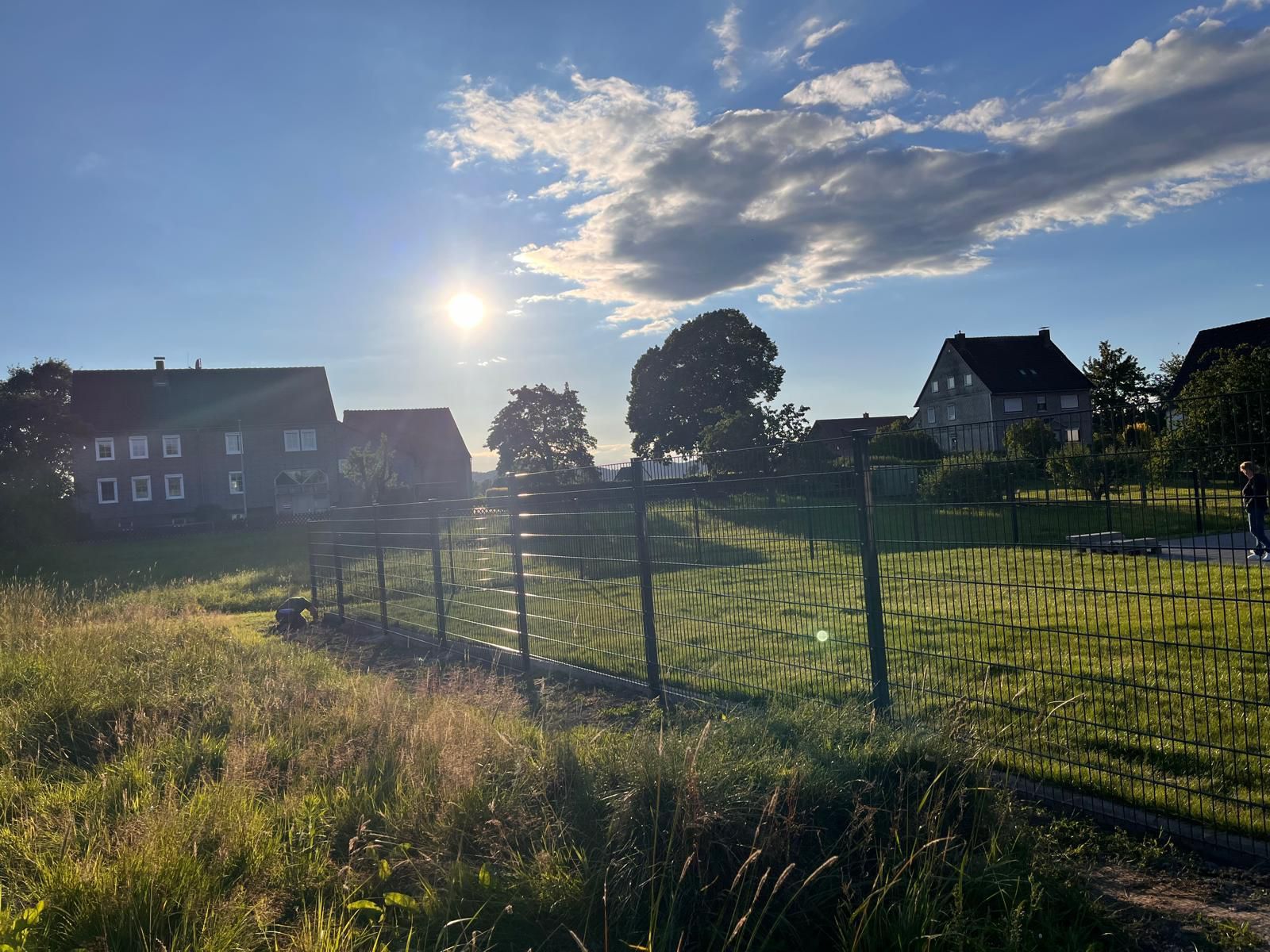 Ein eingezäuntes Feld mit Häusern im Hintergrund und der Sonne, die durch die Wolken scheint.