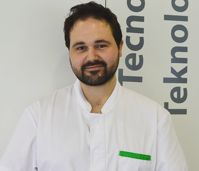 Un hombre con barba y bata blanca de laboratorio se encuentra frente a un cartel que dice tekno