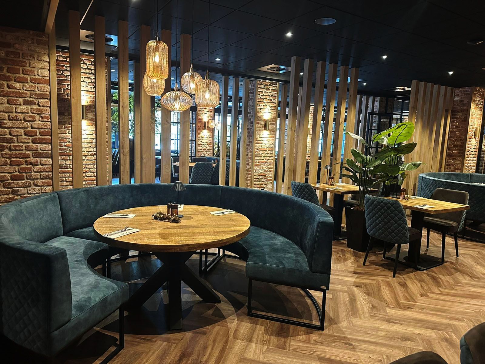 Interieur van een restaurant met een gebogen, blauwgroene bank, houten tafels en decoratieve verlichting.