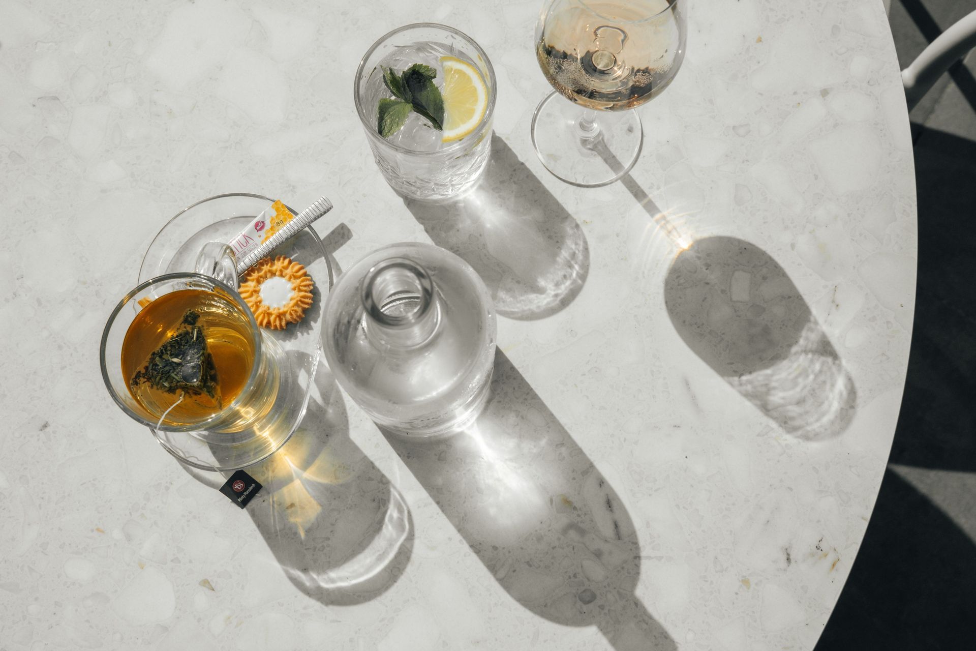 Drankjes op een marmeren tafel die lange schaduwen werpen: thee, een cocktail, bruiswater en een fles.