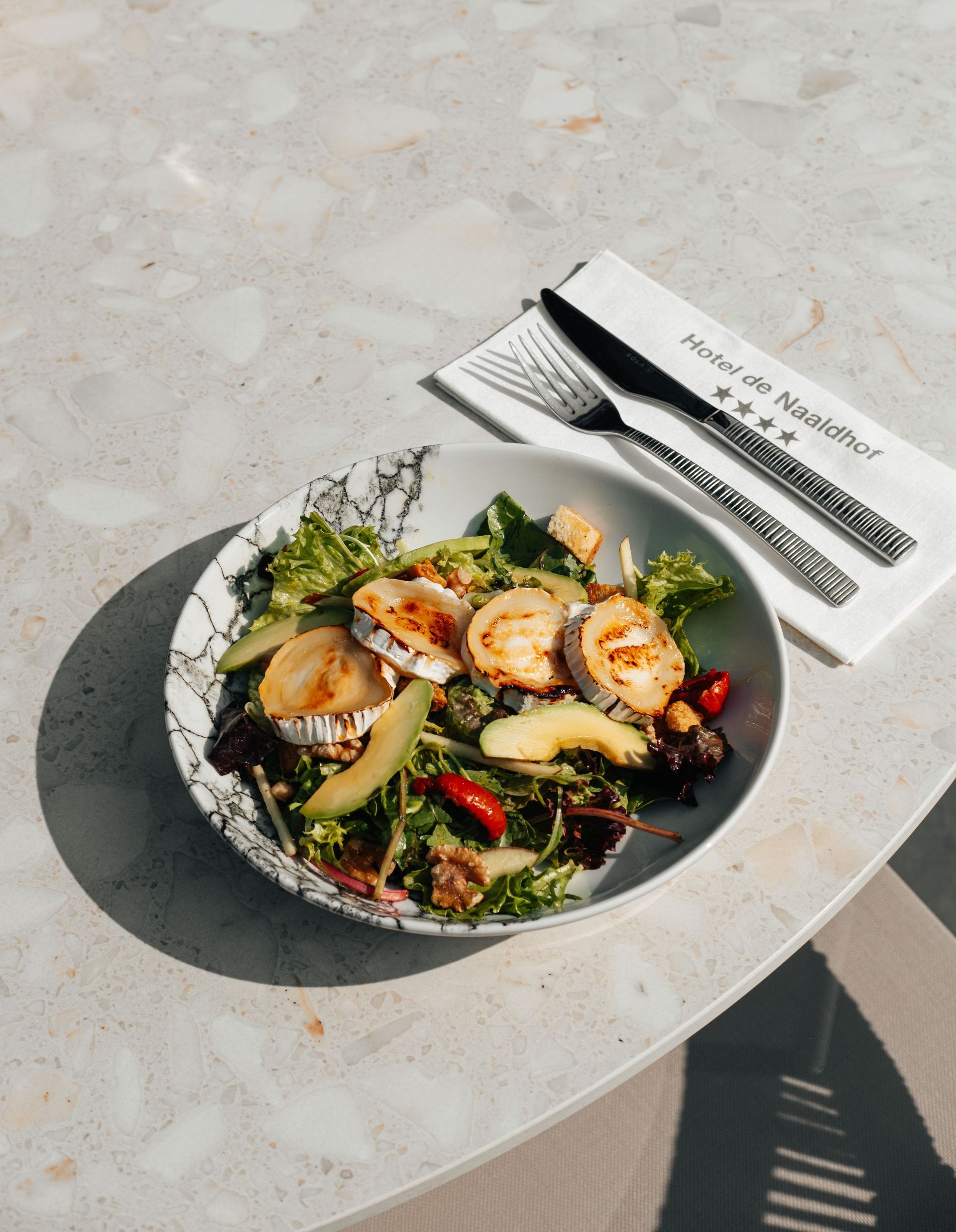 Salade met geitenkaas, avocado en groenten op een bord met patroon, naast bestek op een servet, op een stenen tafel.