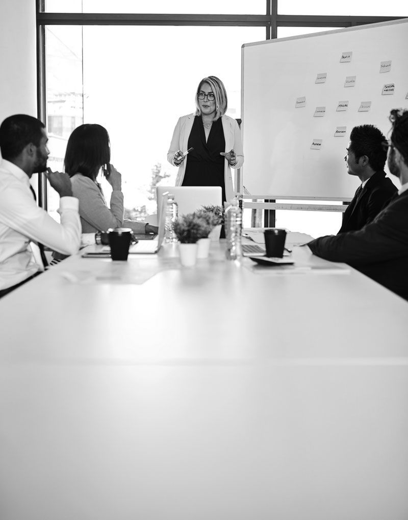 Geschäftstreffen in einem Konferenzraum, mit einem Präsentator an einer Tafel und Teilnehmern, die um einen Tisch sitzen.