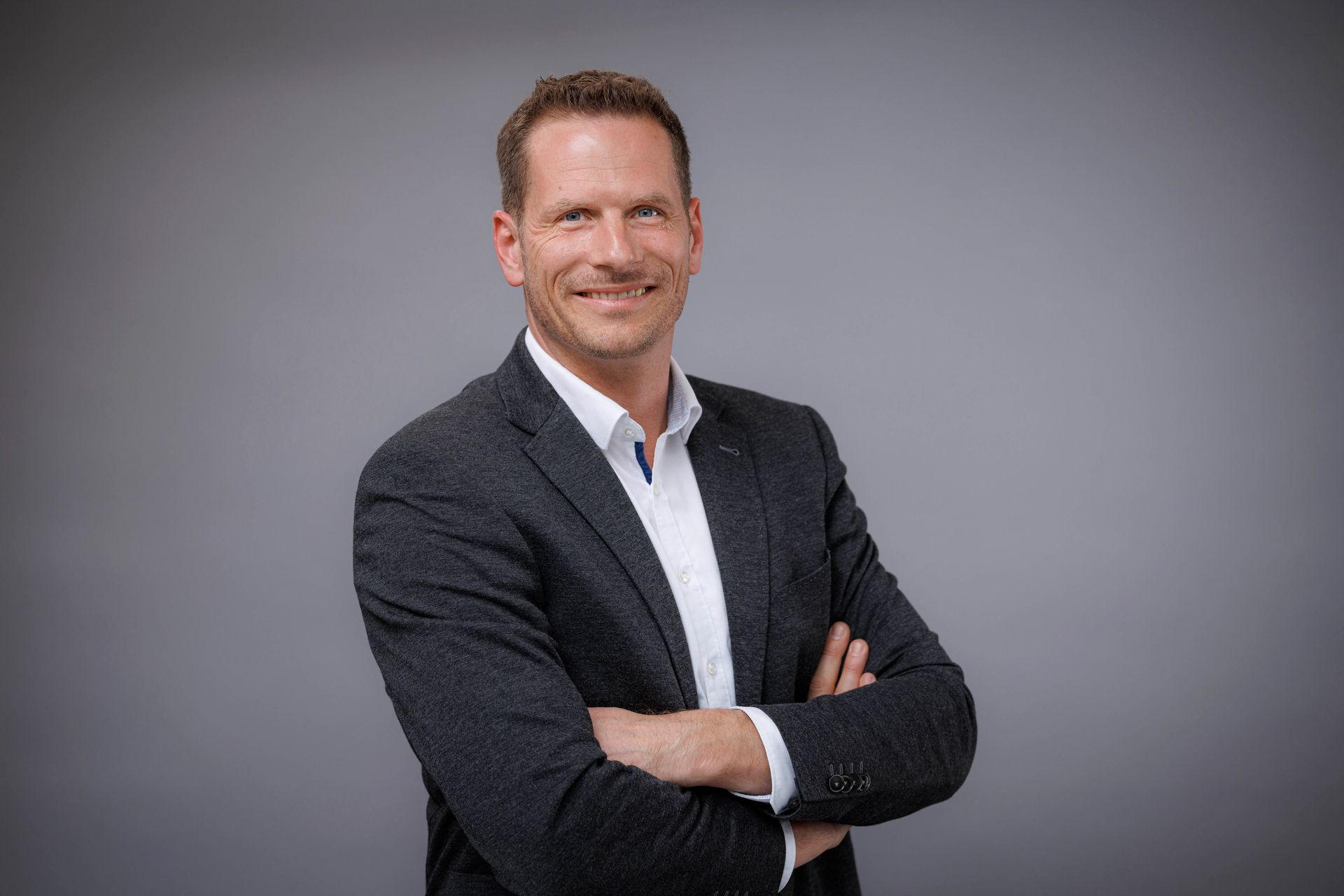 Ein Mann in Anzug und weißem Hemd steht mit verschränkten Armen da und lächelt.
