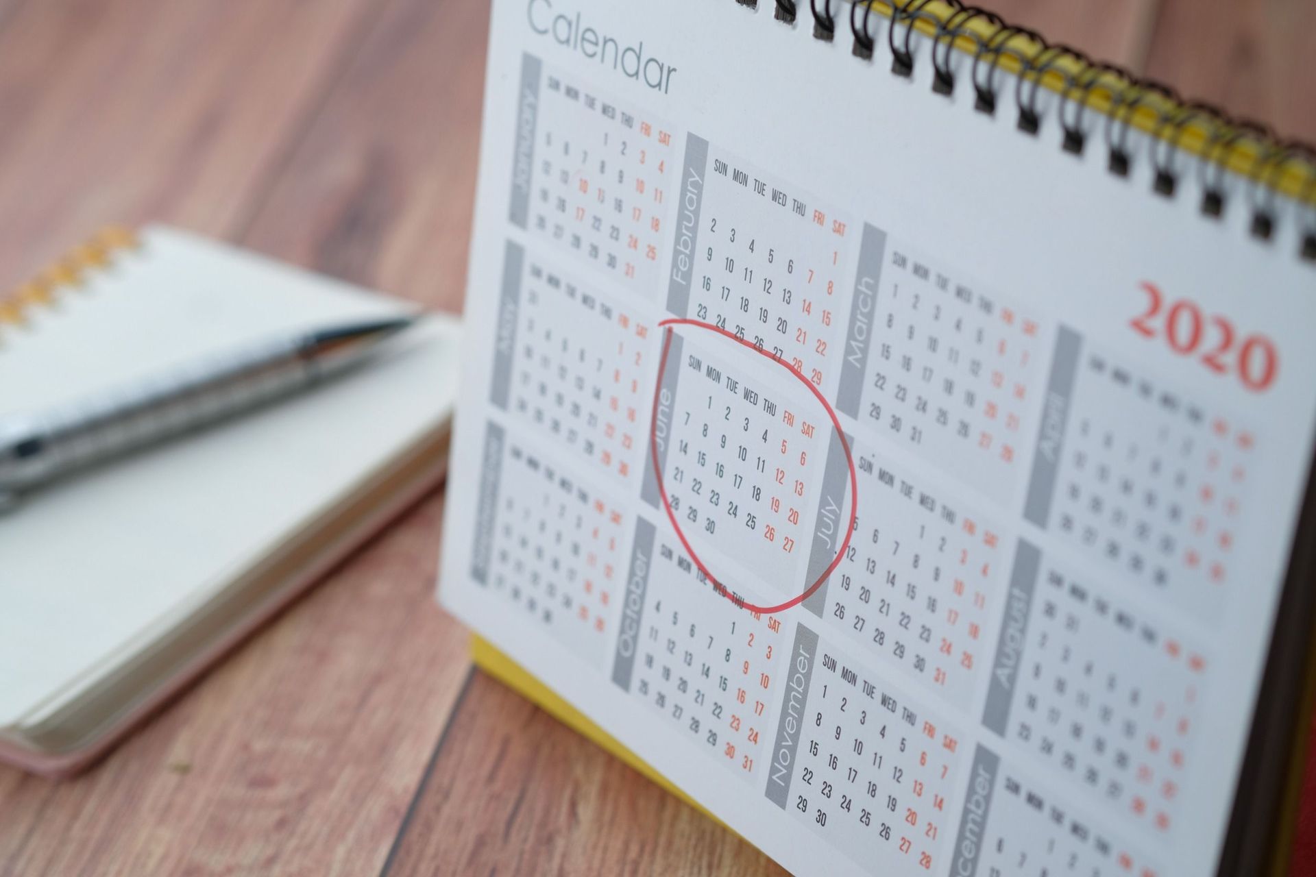 Un calendario para el año 2020 está sobre una mesa de madera junto a un cuaderno y un bolígrafo.