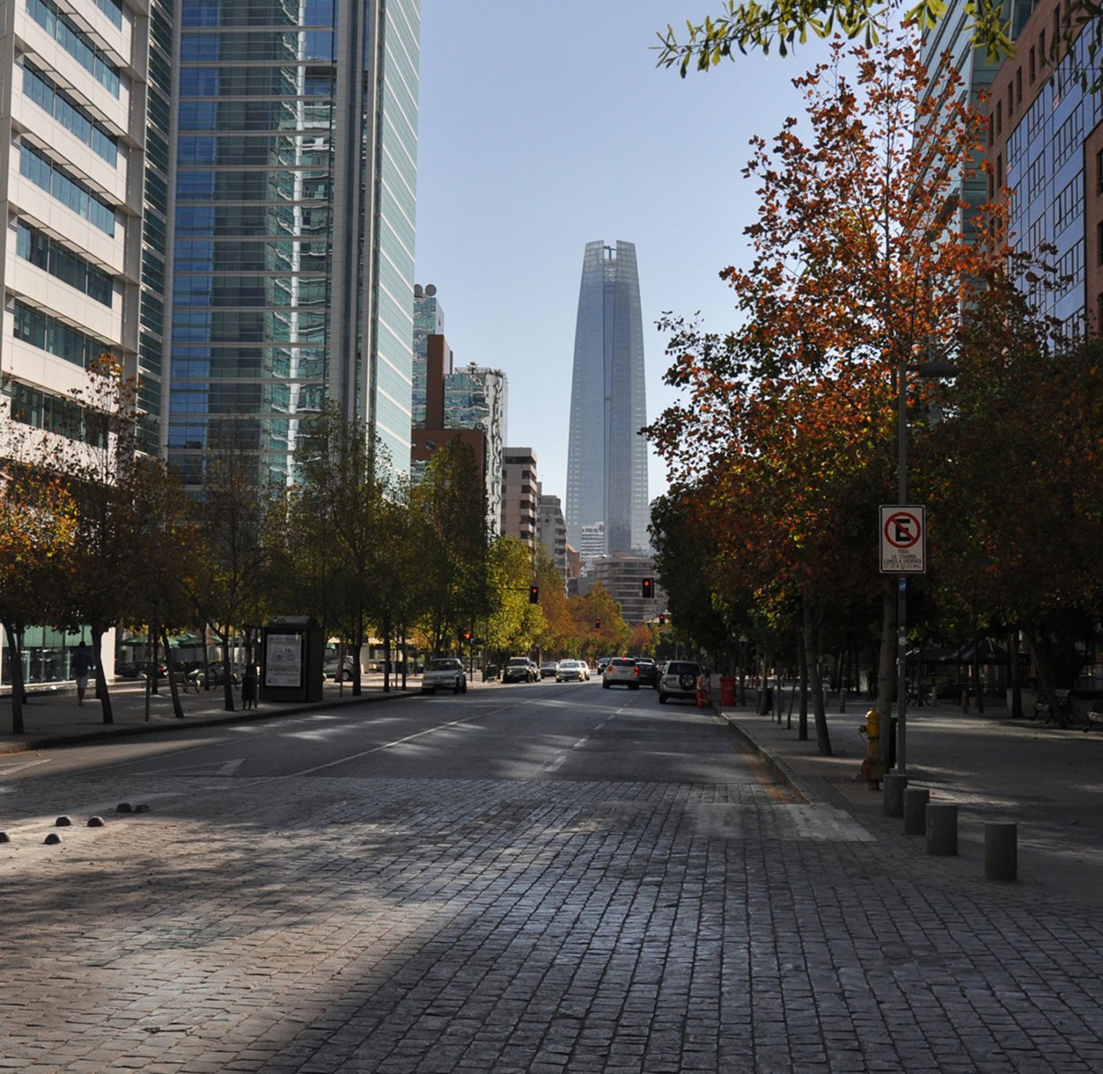Wide city street with trees and tall glass buildings, centered on a distant skyscraper under a clear sky