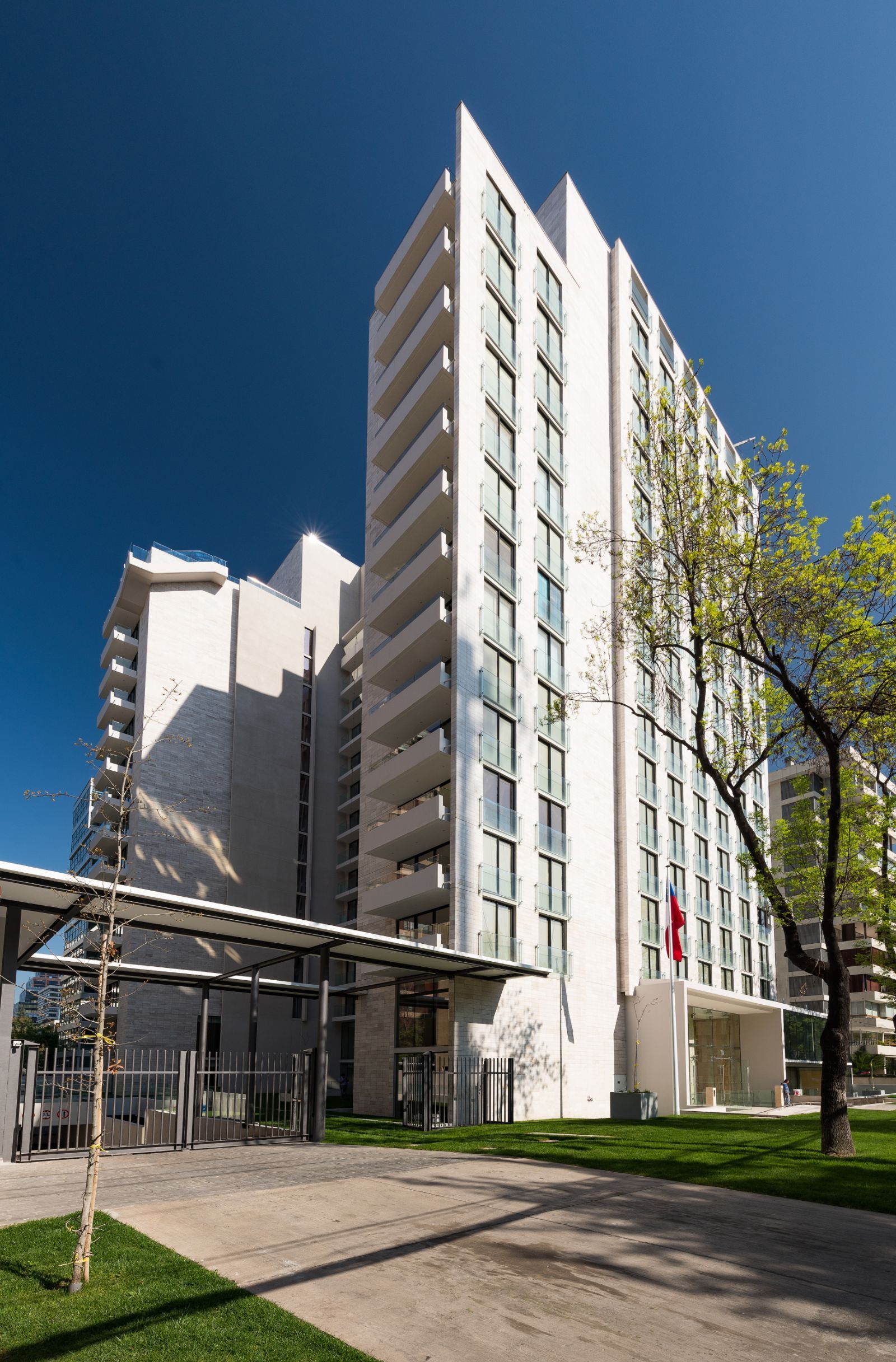 Tall white apartment building with balconies and trees beside a walkway under a clear blue sky