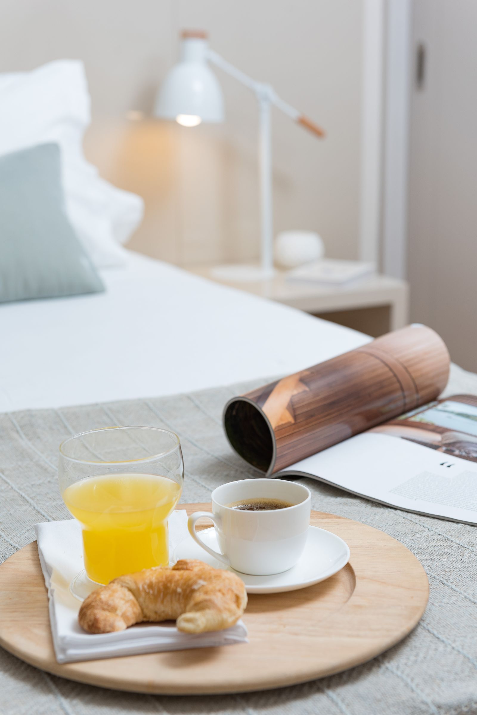 Breakfast tray on a bed with orange juice, coffee, croissant, and a magazine in a bright bedroom
