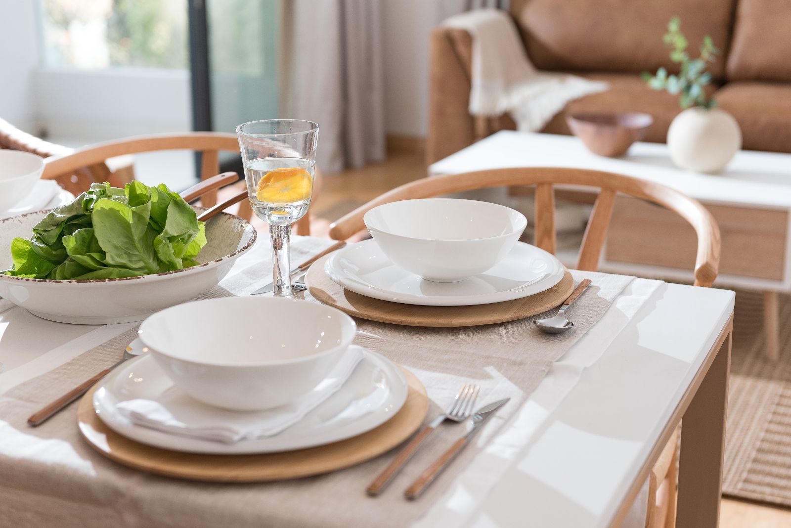 Set dining table with white dishes, woven placemats, and a bowl of green salad by a bright window