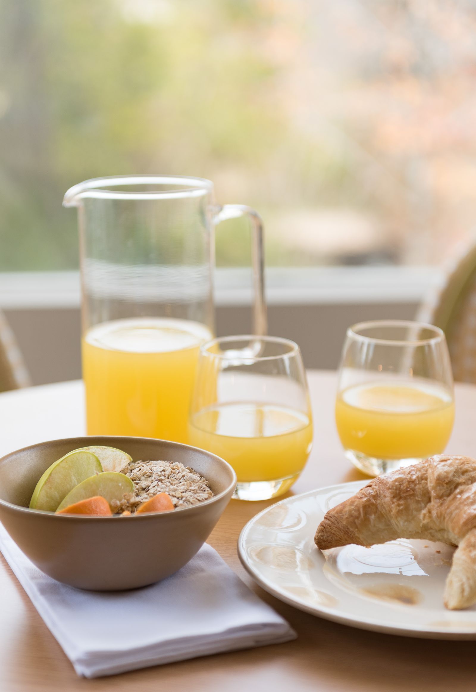 Breakfast table with orange juice, fruit bowl, croissant, and a pitcher by a window