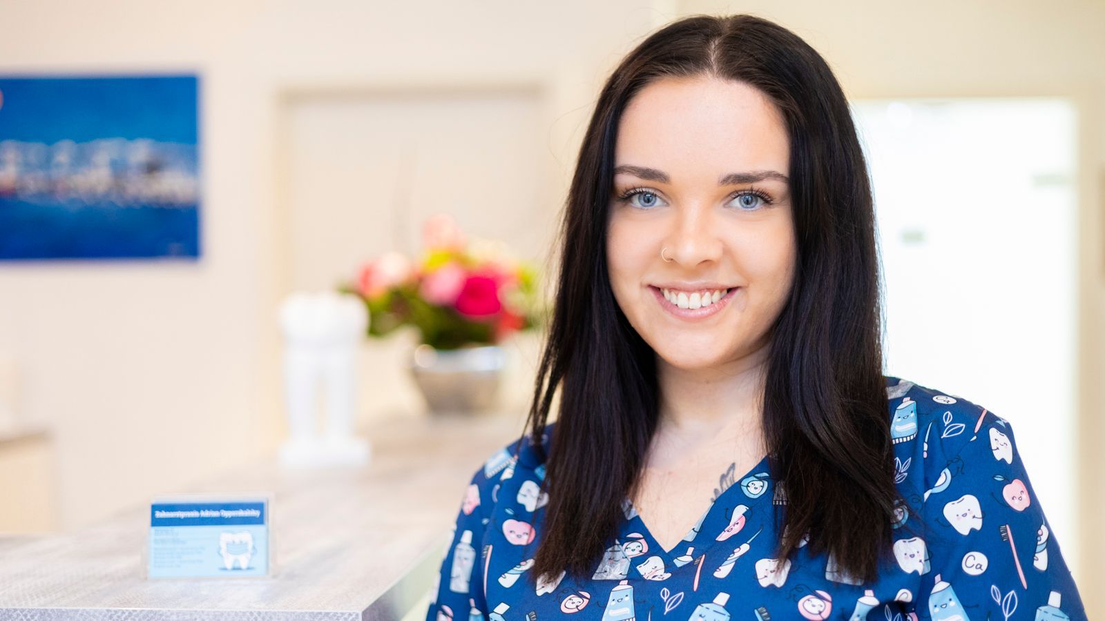 Eine Frau in einem blauen OP-Kittel lächelt in einer Zahnarztpraxis.