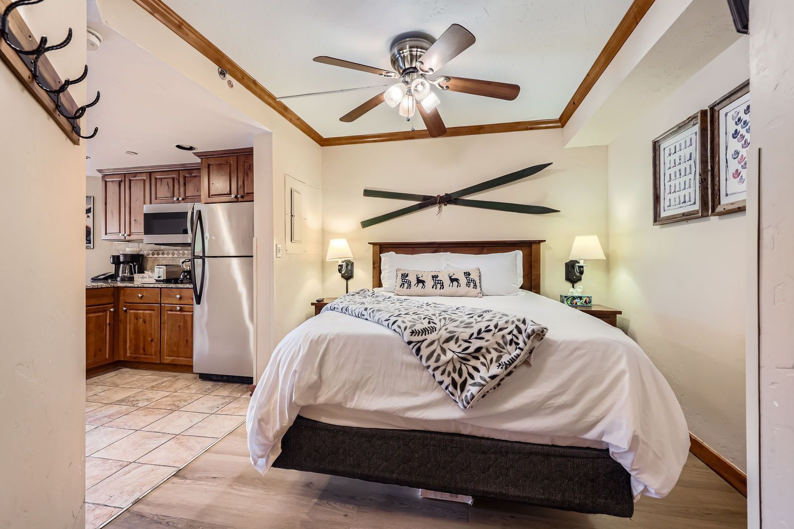 Bedroom with bed, ski decor, and small kitchen. Brown cabinets and a ceiling fan.