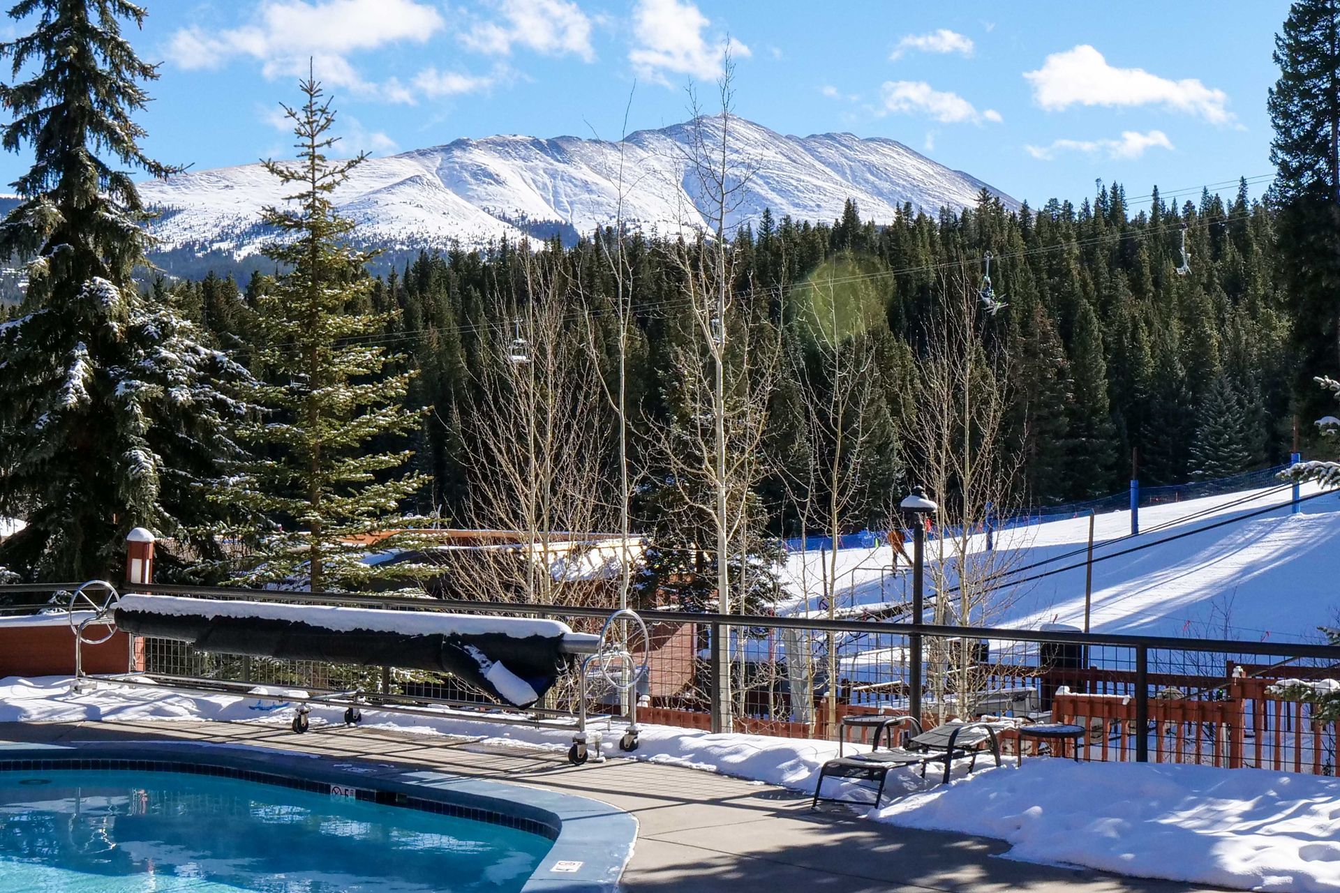 Swimming pool with snow-covered deck, pine trees, and mountain in the background.