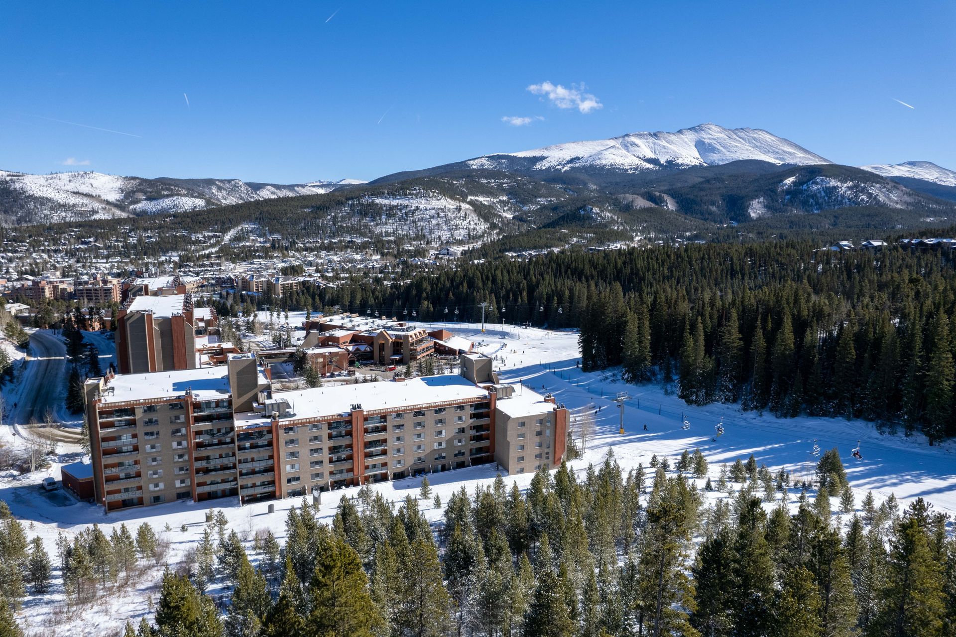 Snow-covered multi-story buildings and trees at a ski resort, with mountains and blue sky in the background.