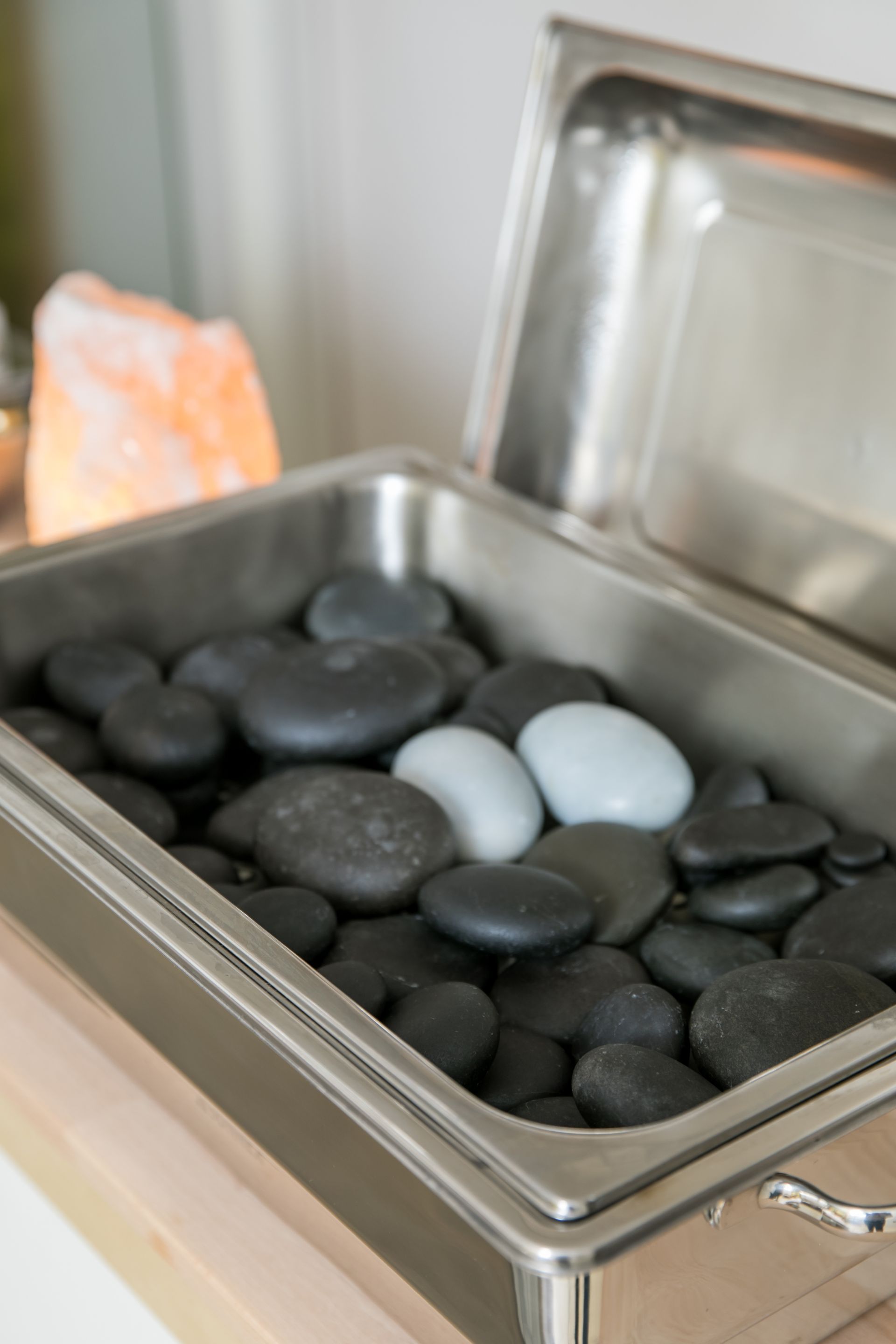 Nahaufnahme eines Metallbehälters gefüllt mit glatten, schwarzen und weißen Massagesteinen, in der Nähe einer brennenden Salzlampe.