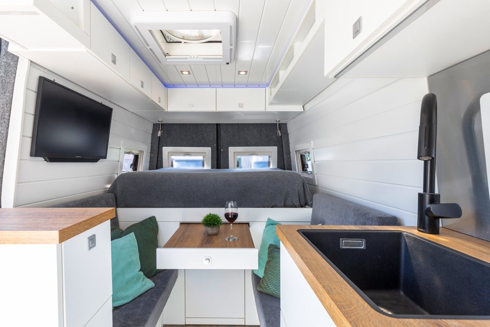 Innenraum eines Wohnmobils mit Bett, Tisch, Spüle und Stauraum über dem Dach, in Weiß-, Grau- und Holztönen.