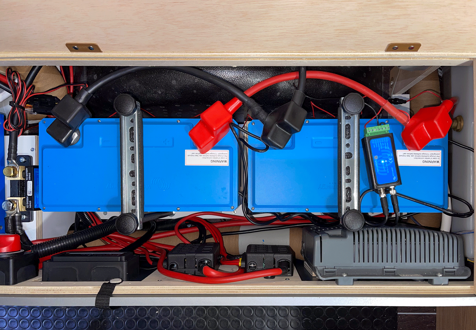 Zwei blaue Batterien in einer Holzbox mit Kabeln, Sicherungen und einem Bildschirm.