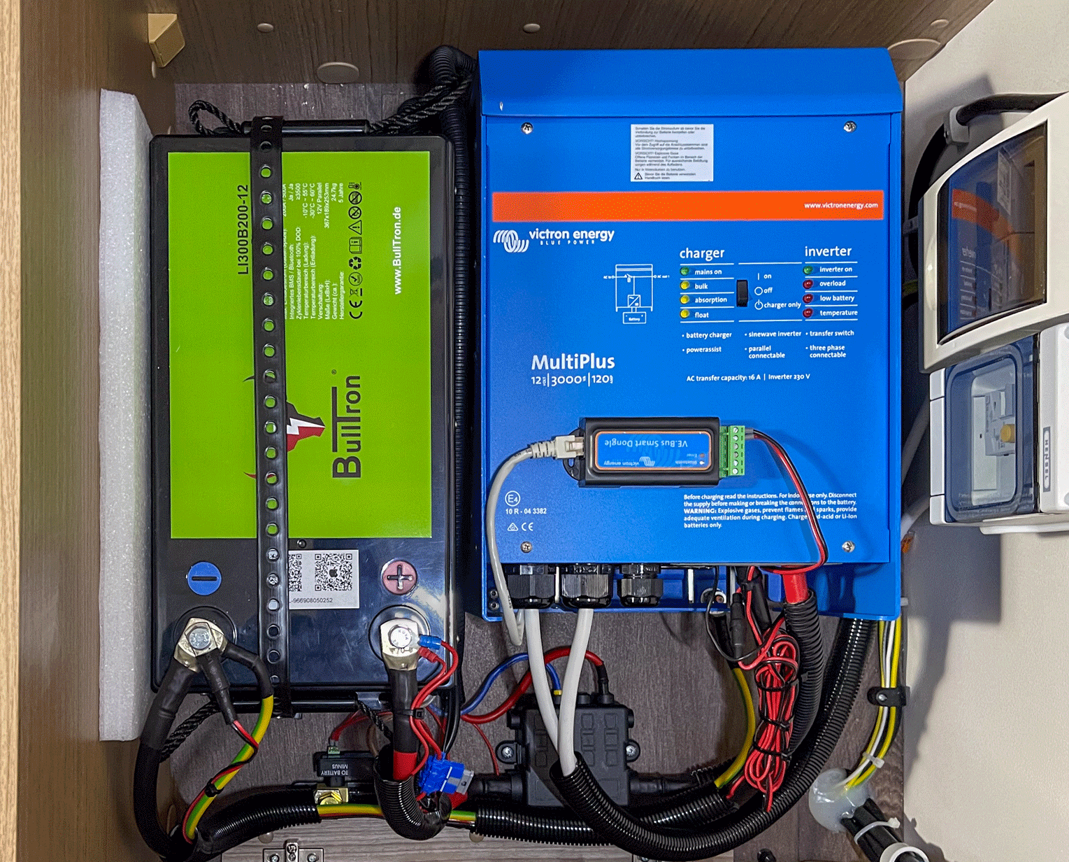 Eine Innenansicht eines Schranks, in dem sich eine grüne Batterie und ein blauer Wechselrichter mit verschiedenen Kabeln und elektrischen Komponenten befinden.