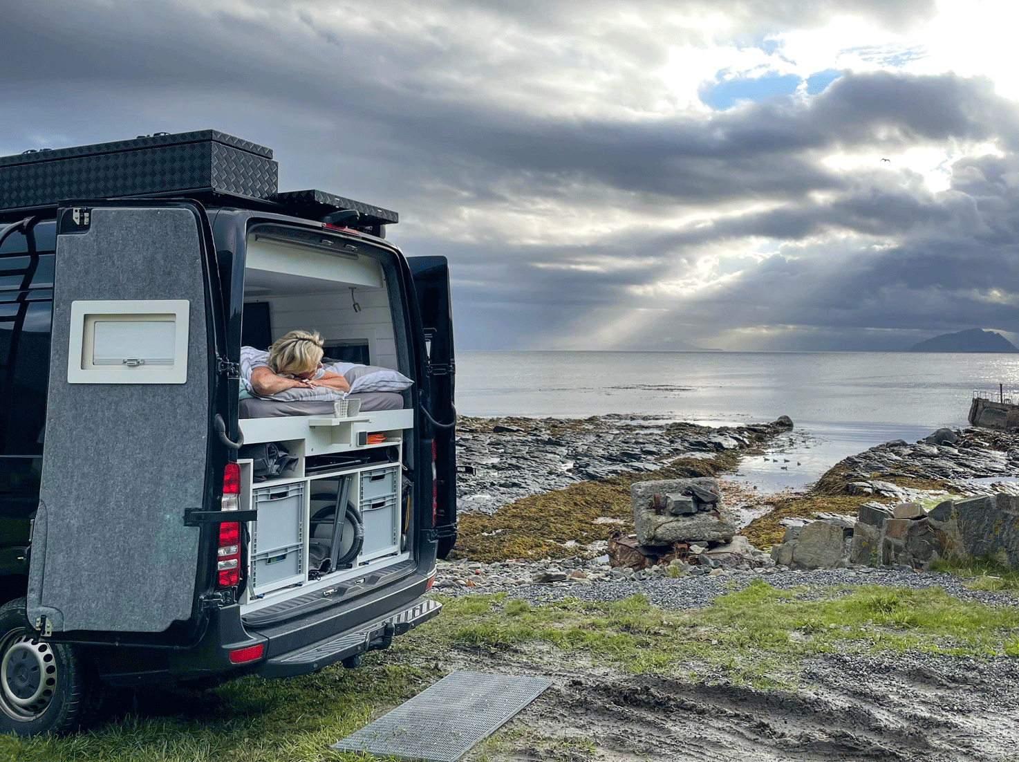 Eine Person auf der Ladefläche eines Lieferwagens mit Blick aufs Meer. Bewölkter Himmel mit Sonnenstrahlen.