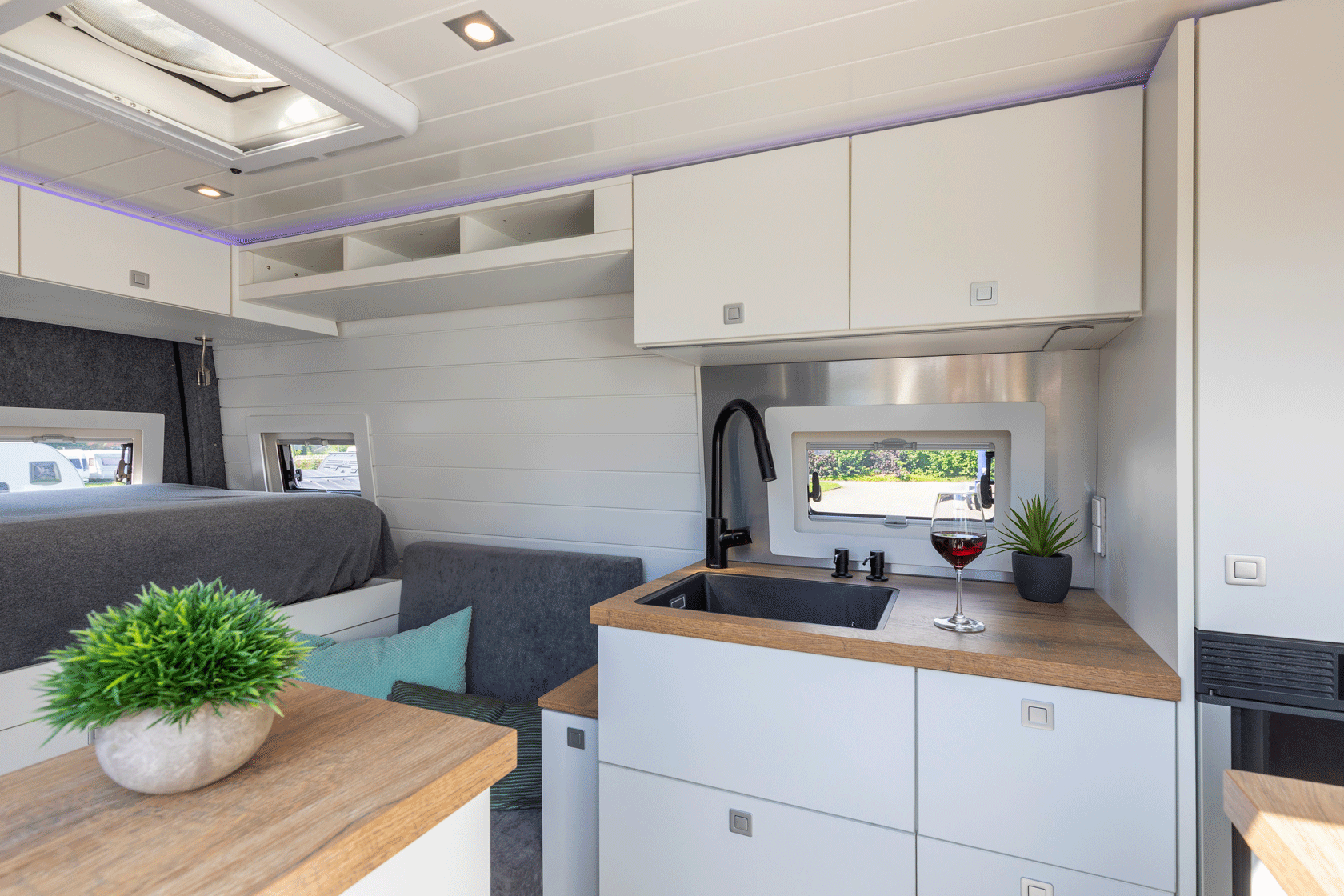 Innenraum einer modernen Wohnmobilküche mit weißen Schränken, kleiner Spüle und einem Sofa.