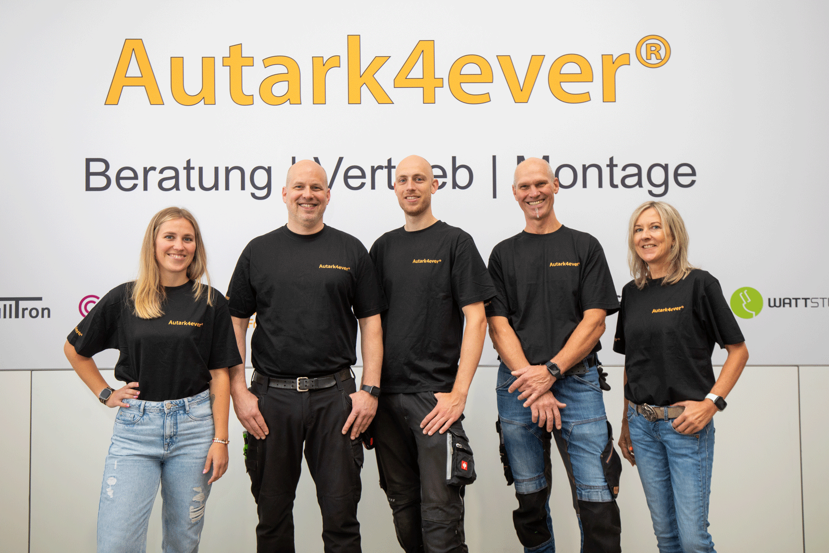 Fünf Personen in schwarzen T-Shirts mit dem Logo „Autark4ever“ posieren vor einer weißen Wand, vermutlich ein Firmenfoto.