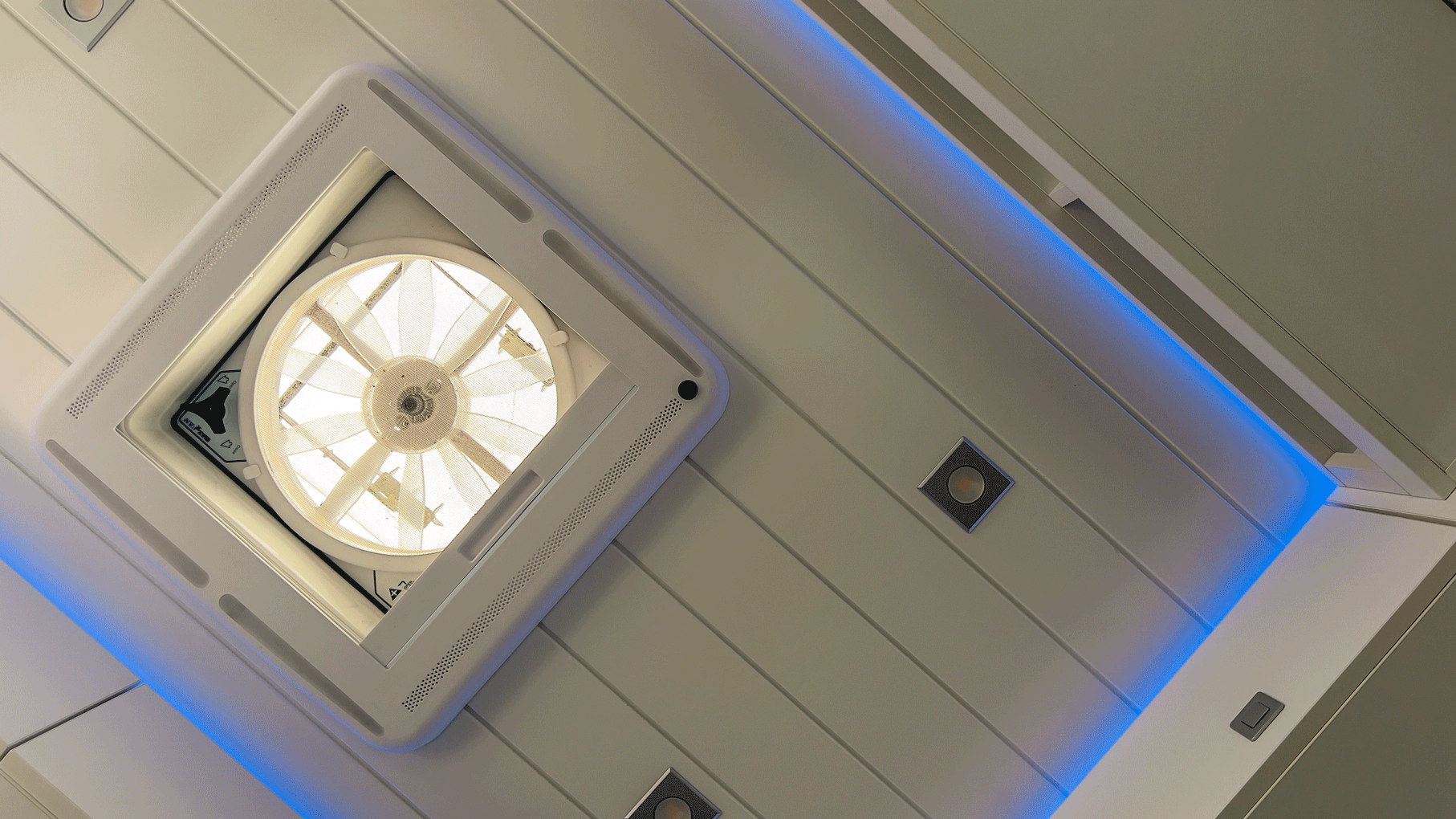 Deckenventilator mit weißem Rahmen und Flügeln, umgeben von weißer Verkleidung und blauen Akzentlichtern.