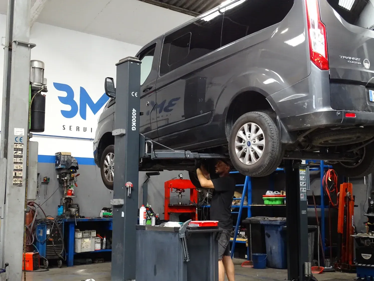Un mecánico trabajando debajo de una furgoneta gris elevada en un elevador dentro de un taller de reparación.