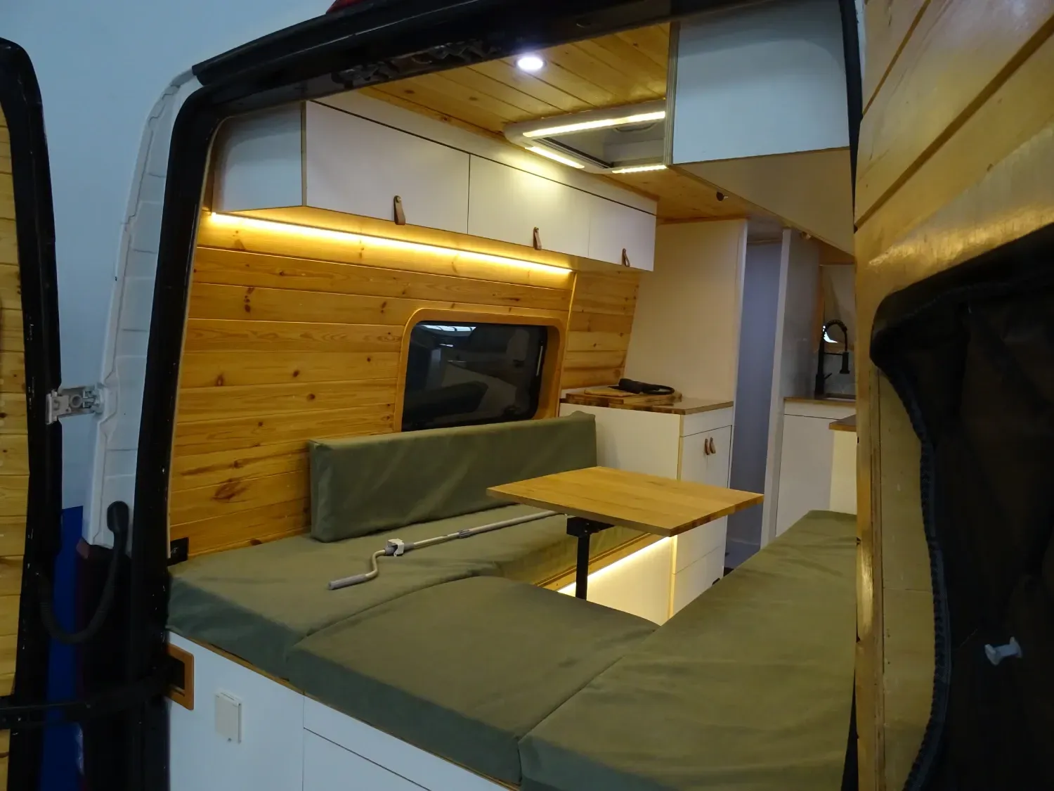 Vista interior de una furgoneta transformada con zona de asientos tipo banco, detalles de madera y armarios superiores.