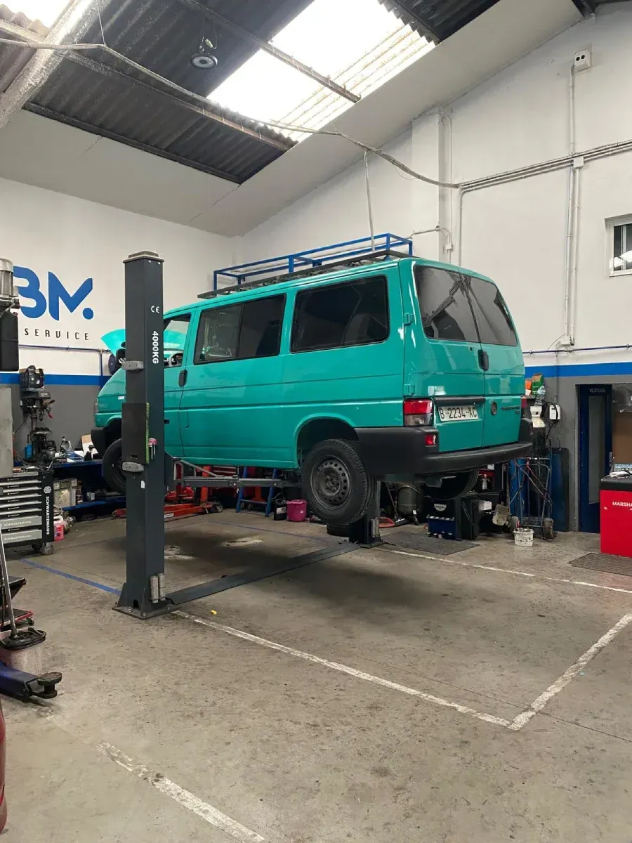 Furgoneta color verde azulado sobre un elevador en un garaje, con la baca visible.