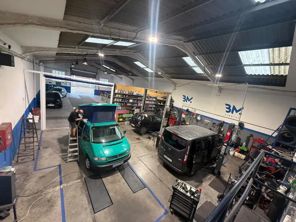 Interior de un taller con una furgoneta verde y una furgoneta gris. Hay trabajadores presentes y herramientas a la vista.