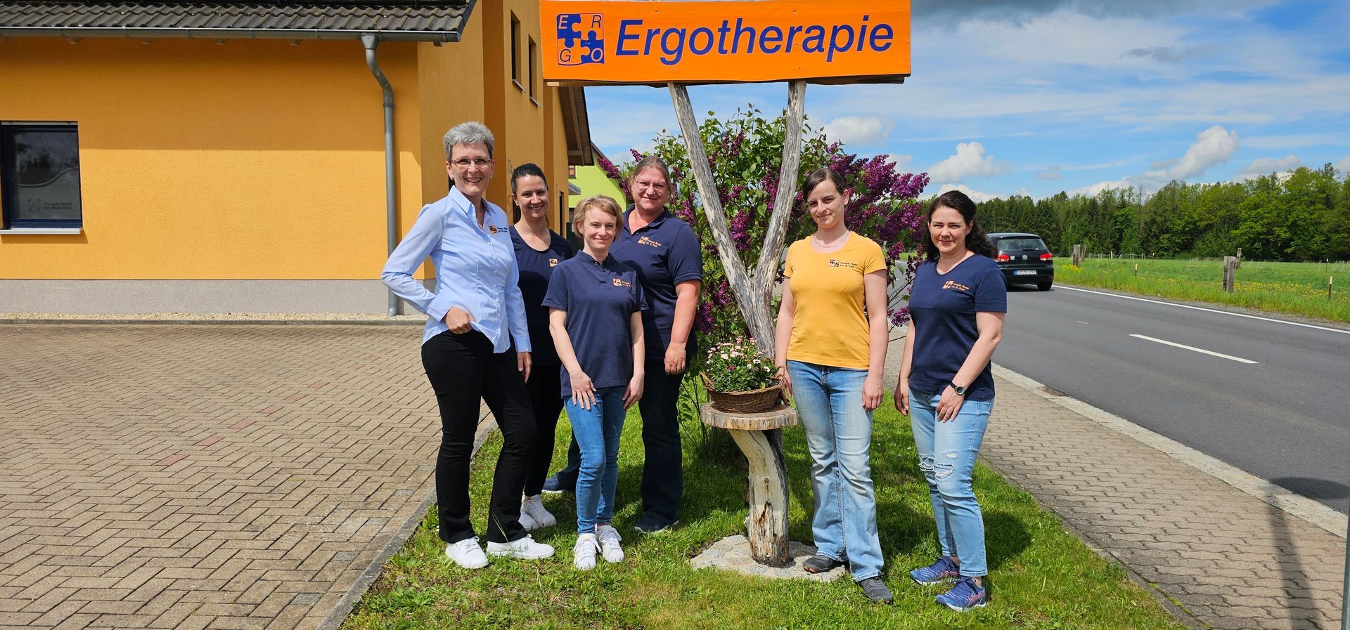 Fünf Personen stehen an einem sonnigen Tag im Freien neben einem Schild einer Ergotherapiepraxis.