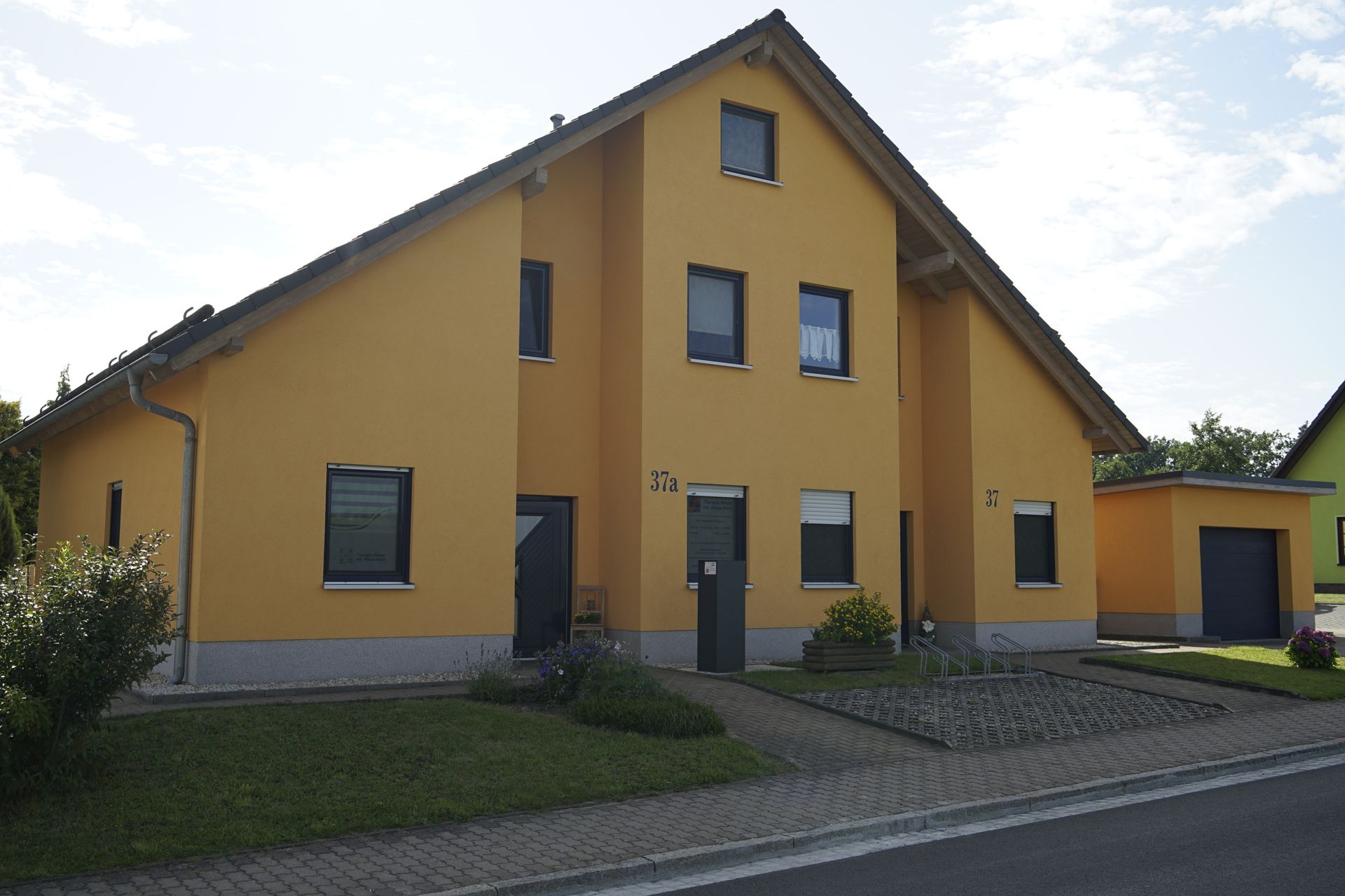 Gelbes zweistöckiges Haus mit dunklen Türen und Fenstern; kleine Garage auf der rechten Seite.