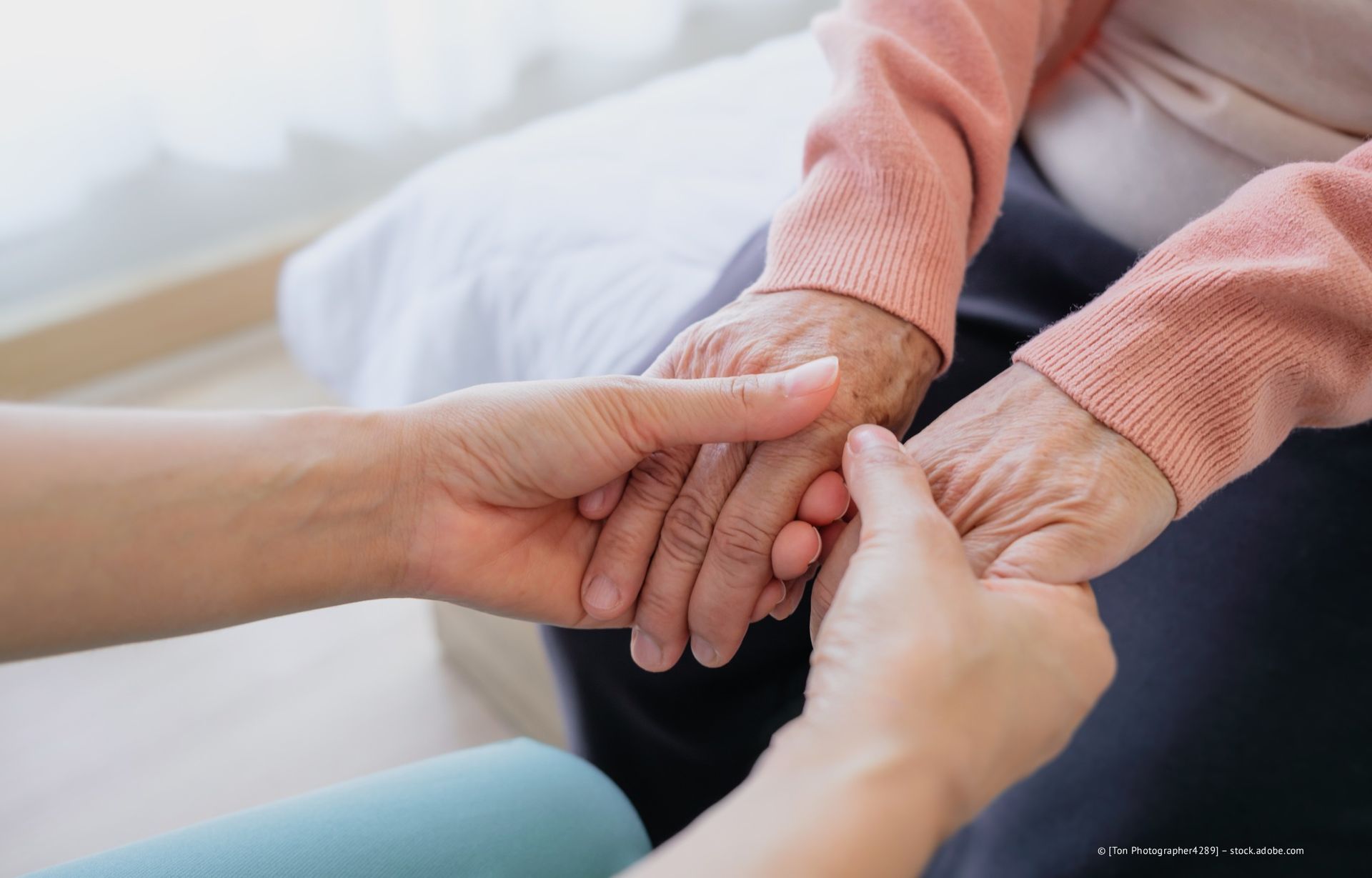 Hände einer älteren Person werden von einer anderen gehalten und bieten Unterstützung an. Helle Haut, rosa Pullover, drinnen.