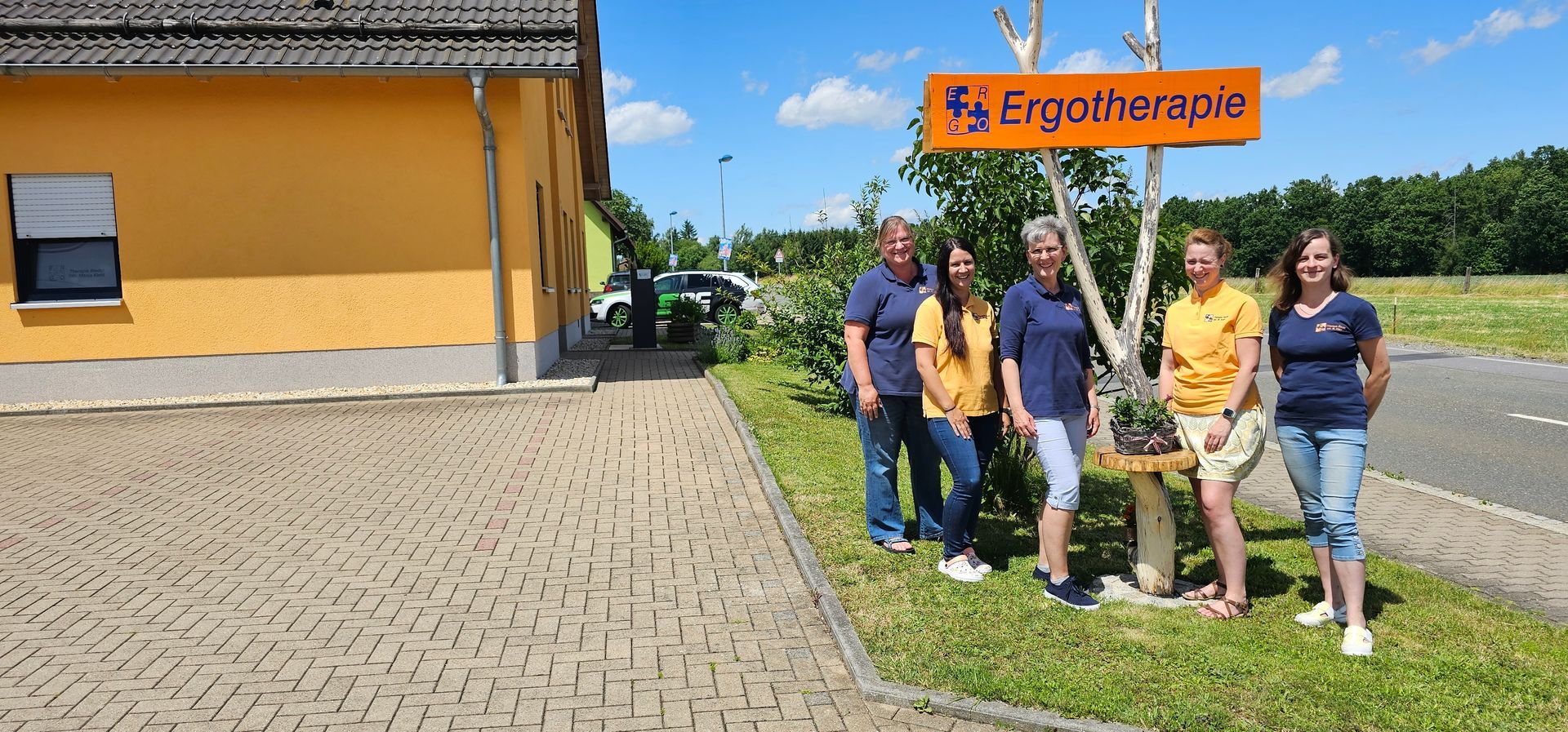 Fünf Frauen stehen neben einem orangefarbenen „Ergotherapie“-Schild. Die Szene scheint im Freien an einem sonnigen Tag zu sein.