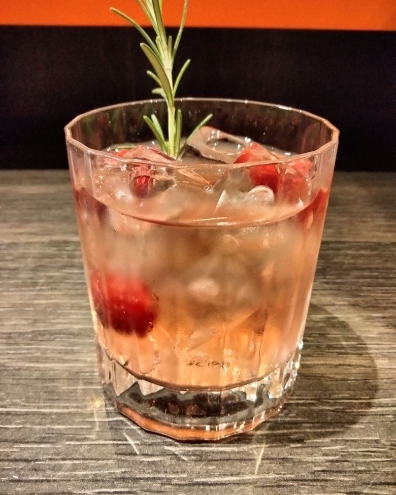 Un cocktail servi dans un verre avec des glaçons, des canneberges entières et un brin de romarin.