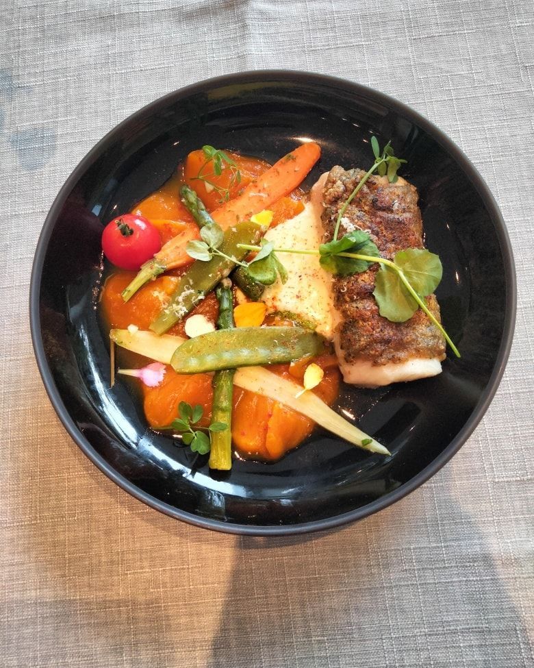 Un plat gastronomique servi dans un bol noir, composé de poisson poêlé, de légumes rôtis colorés.