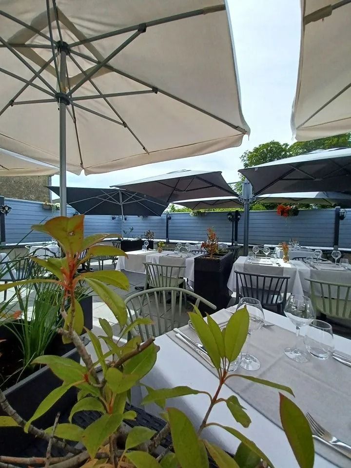 Terrasse extérieure avec tables dressées pour un repas sous de grands parasols, avec de la verdure au premier plan.