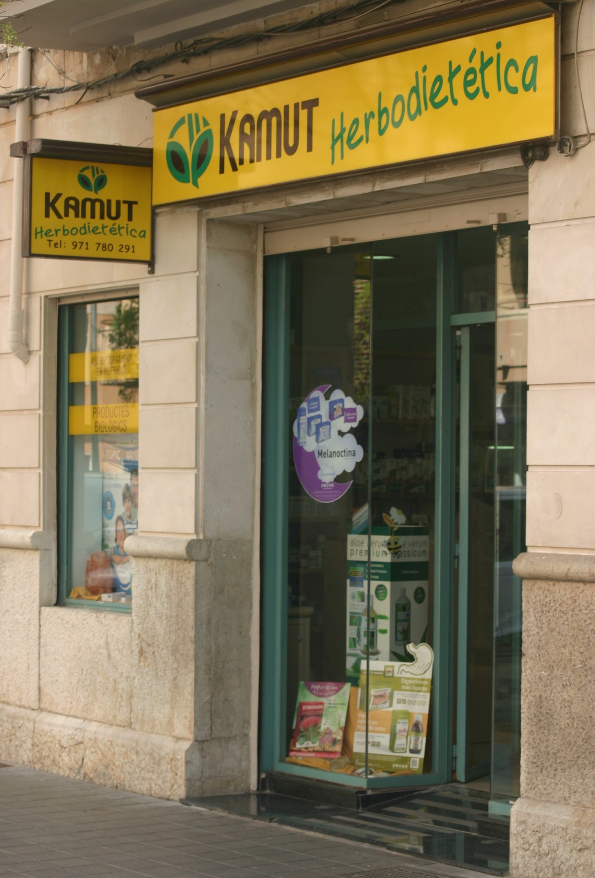 Fachada de KAMUT Herbodietética con letrero amarillo y entrada de cristal en un edificio de piedra.