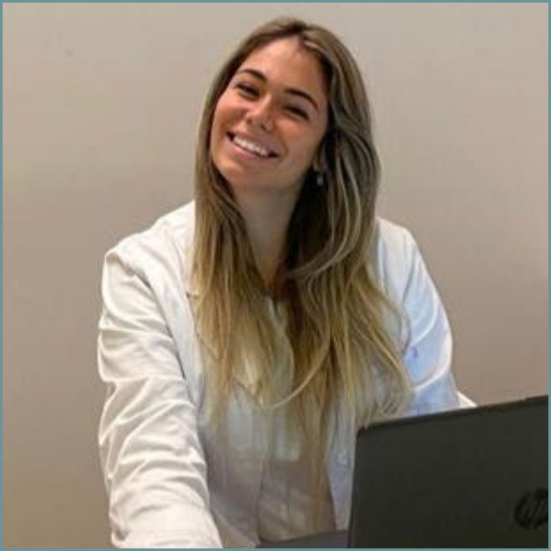 Una giovane donna con i capelli lunghi e biondi  in camice bianco sorride, seduta alla scrivania con un computer portatile.