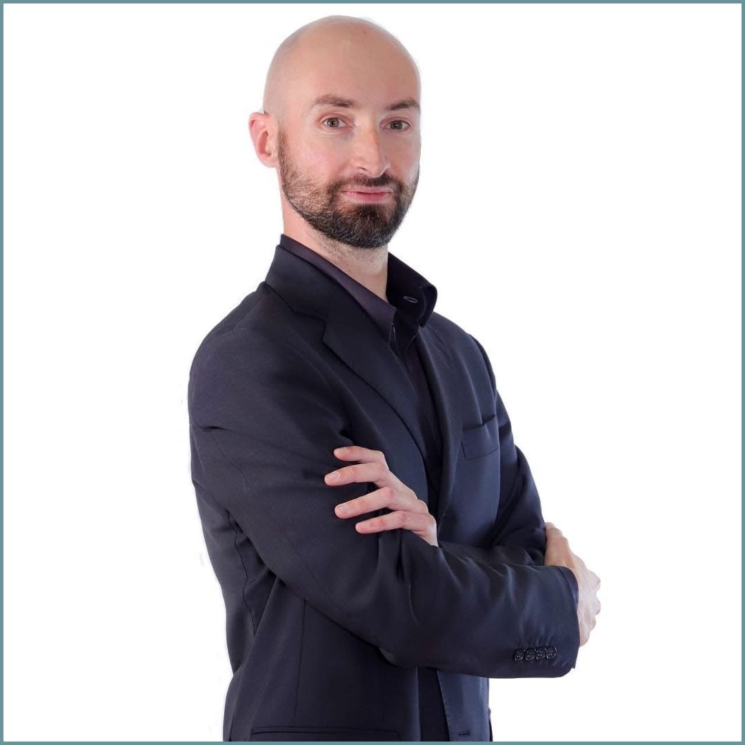 Uomo calvo con barba curata, braccia incrociate, vestito con un abito scuro e una camicia con il colletto aperto.