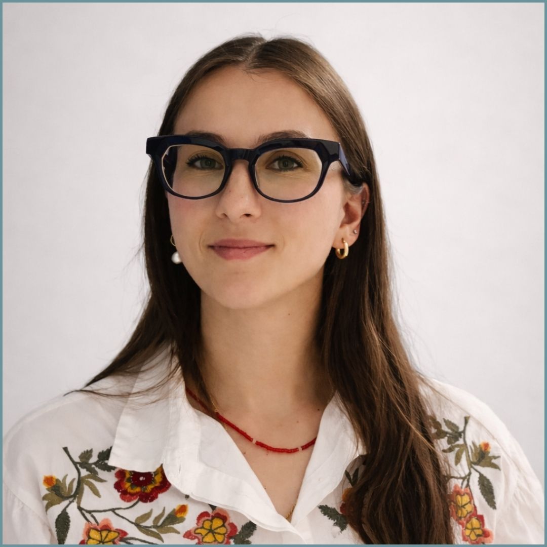 Una giovane donna con lunghi capelli castani sorride, indossa una camicetta bianca con un motivo fiorato e occhiali con montatura scura ed è rivolta verso la telecamera.