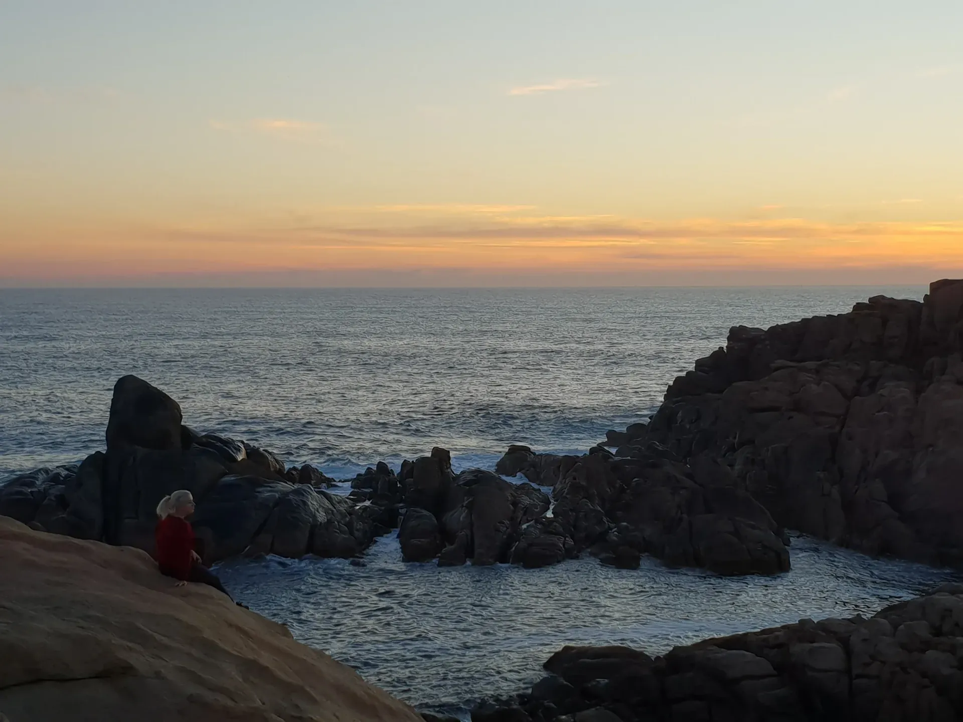 Una persona está sentada en una roca mirando el océano al atardecer.
