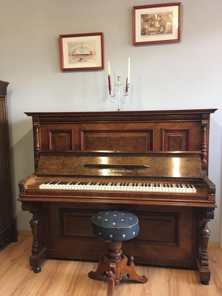 Ein hölzernes Klavier mit Hocker, gerahmte Kunstwerke und Kerzenständer vor einer grauen Wand.