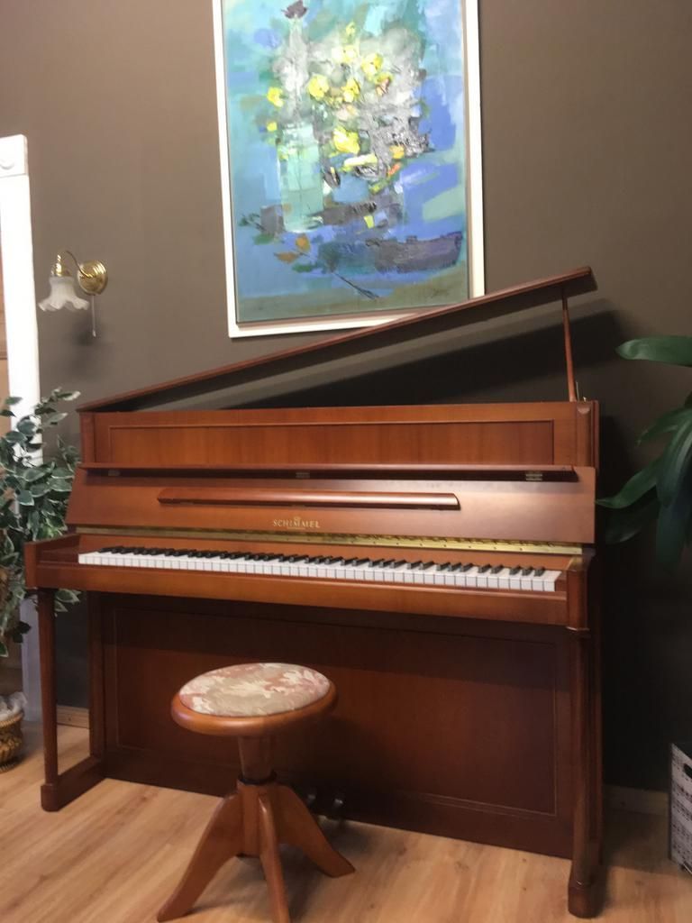 Ein hölzernes Klavier mit geöffnetem Deckel, ein Hocker und ein Kunstwerk an einer braunen Wand.