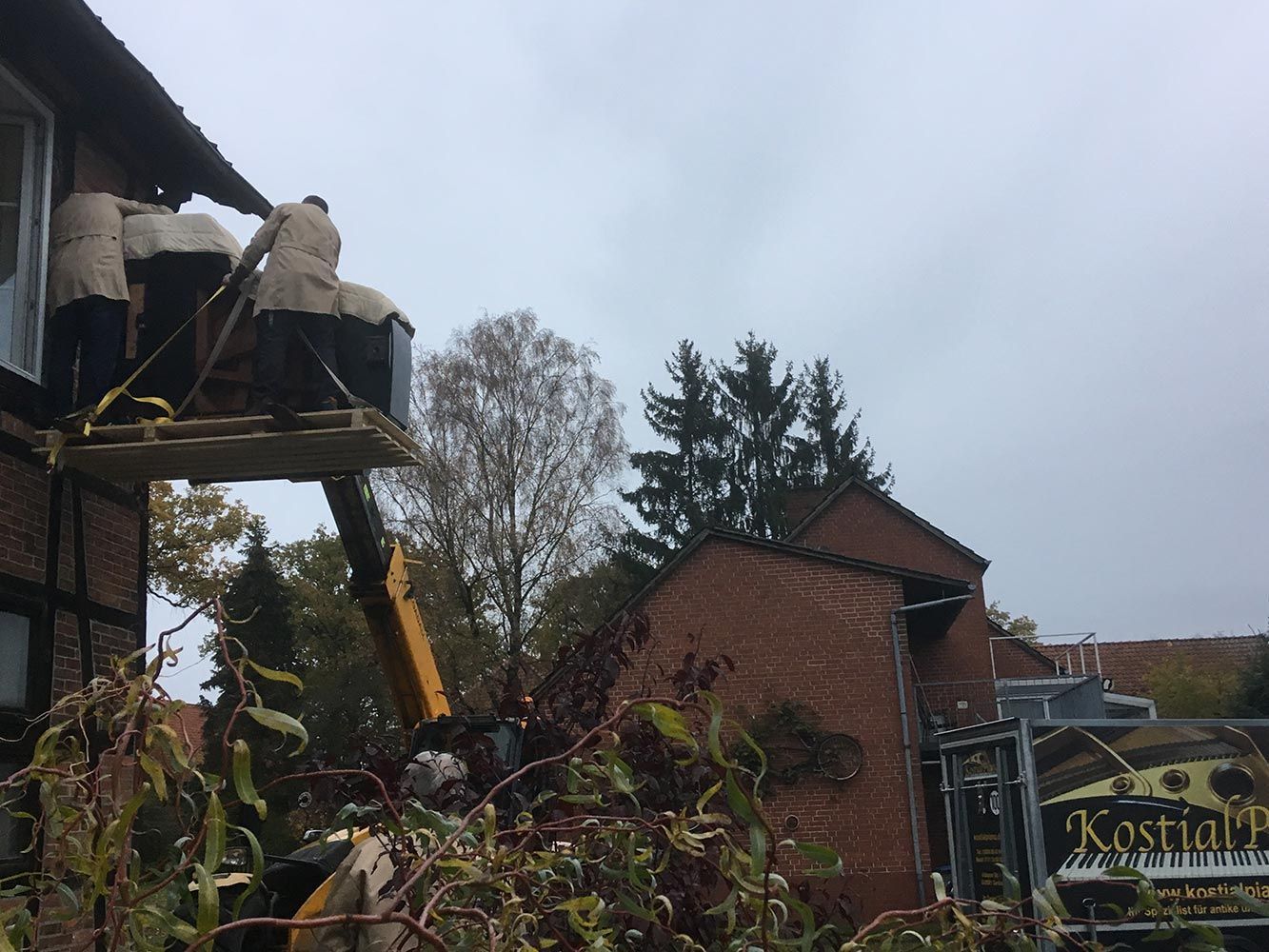 Zwei Personen verladen große Container mit einem Hubwagen auf das Dach eines Gebäudes. Backsteingebäude unter bewölktem Himmel.