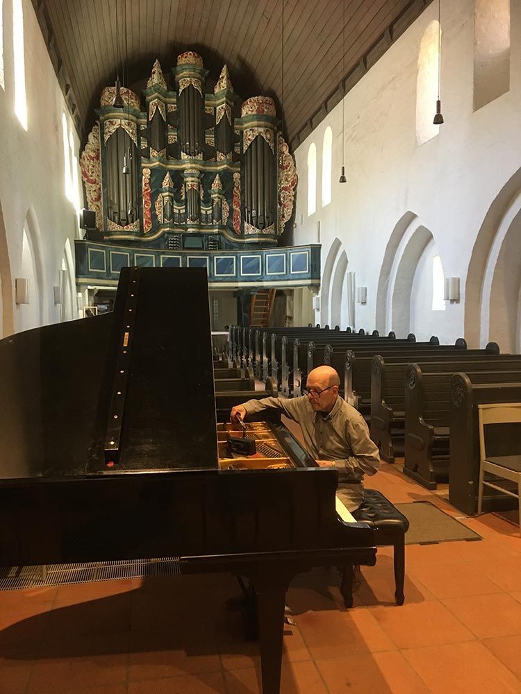 Ein Mann sitzt in einer Kirche an einem Klavier und bereitet sich aufs Spielen vor. Im Hintergrund ist eine Orgel zu hören.