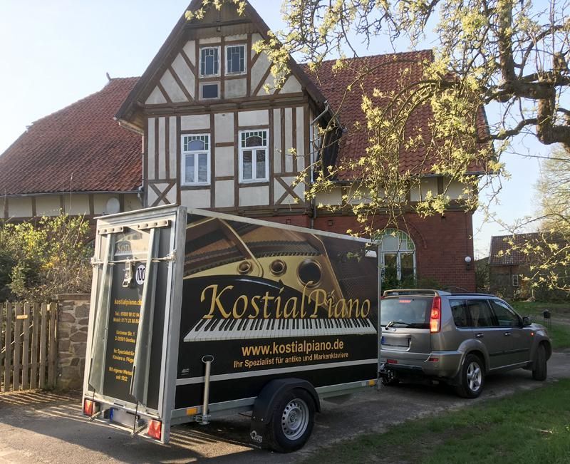 Ein Klaviertransportanhänger steht vor einem Fachwerkhaus; ein Auto ist an den Anhänger angehängt.