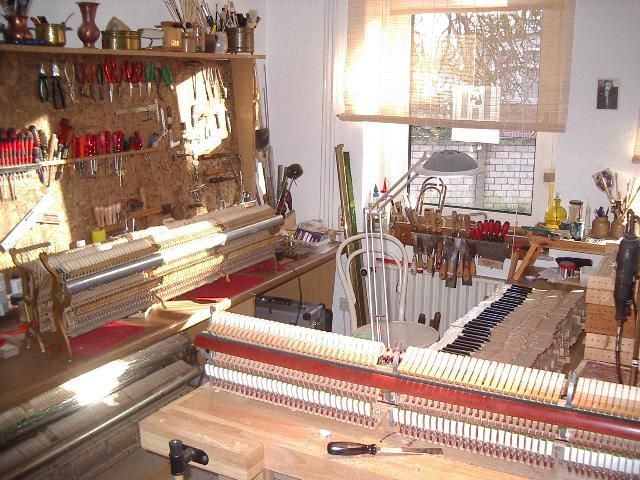 Klavierreparaturwerkstatt. Werkbänke mit freiliegender Klaviermechanik, Werkzeuge in Regalen, natürliches Licht durchs Fenster.