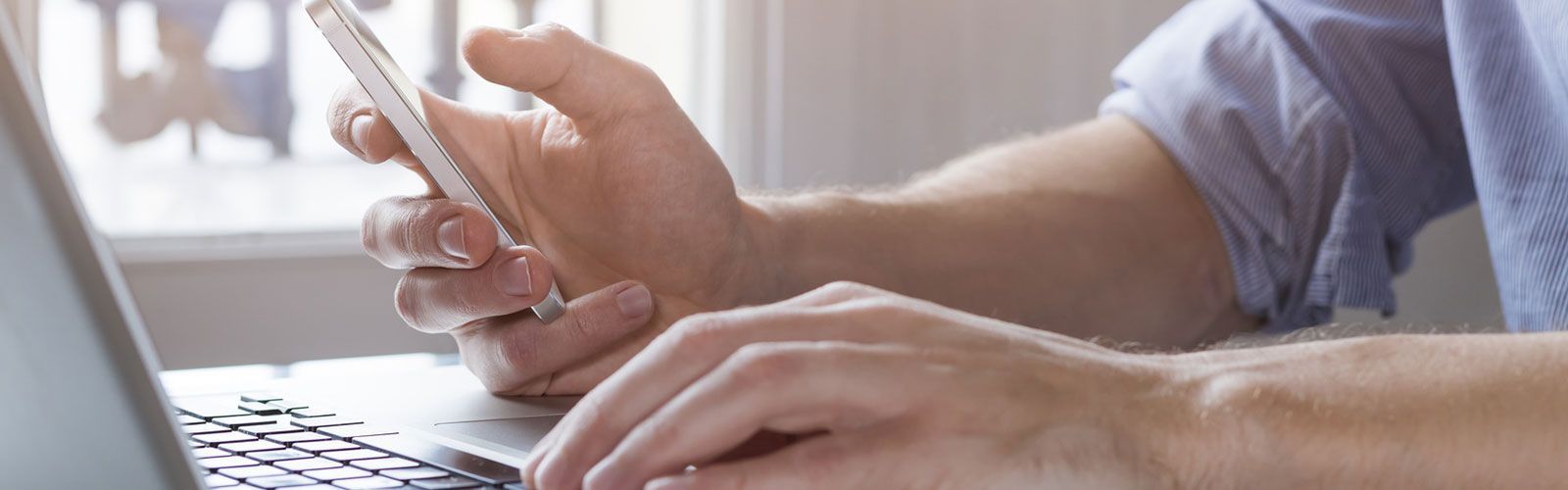 Ein Mann tippt auf einem Laptop und hält dabei ein Mobiltelefon in der Hand.