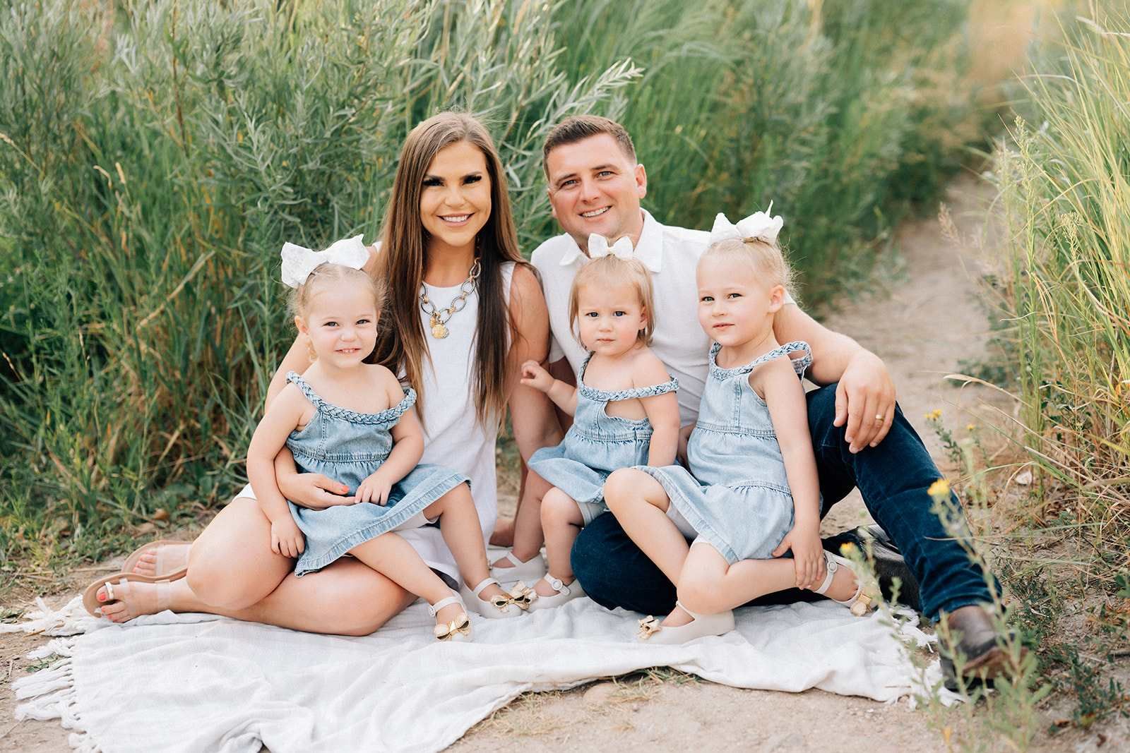 Family of five sitting outdoors on a blanket; mom, dad, and three young children, all smiling.